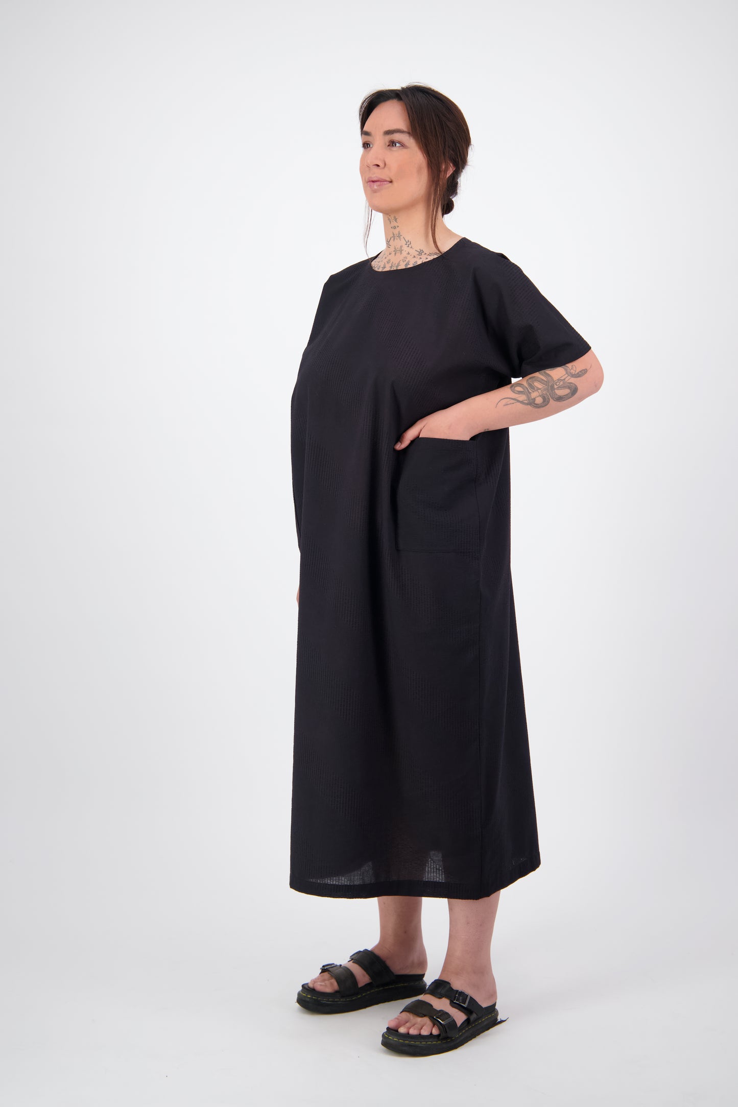 Arlena standing in long black dress with short sleeves and pockets. light seersucker cotton fabric