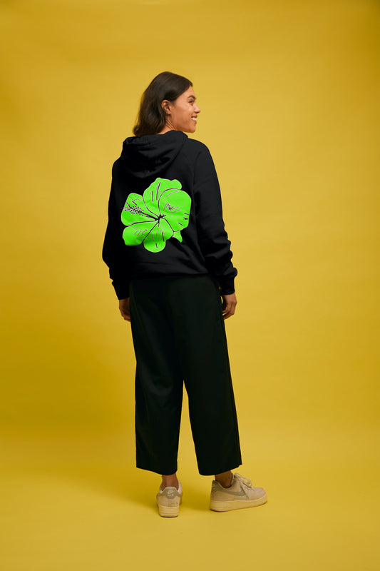 Karen Valerie standing in front of yellow wall wearing black AS colour hoodie with green PAPA text and green hibiscus flower with Navy Buffet pants