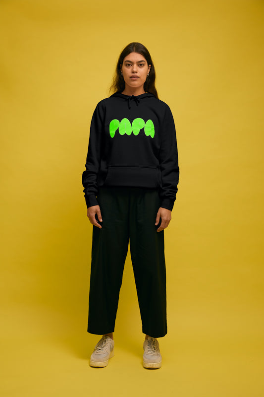 Karen Valerie standing in front of yellow wall wearing black AS colour hoodie with green PAPA text and Navy Buffet pants