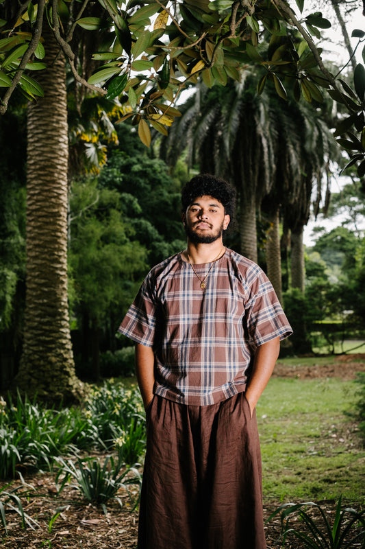 Pacific artist, Keciano wearing a short sleeve brown plaid check t-shirt tee and brown linen pants in a park
