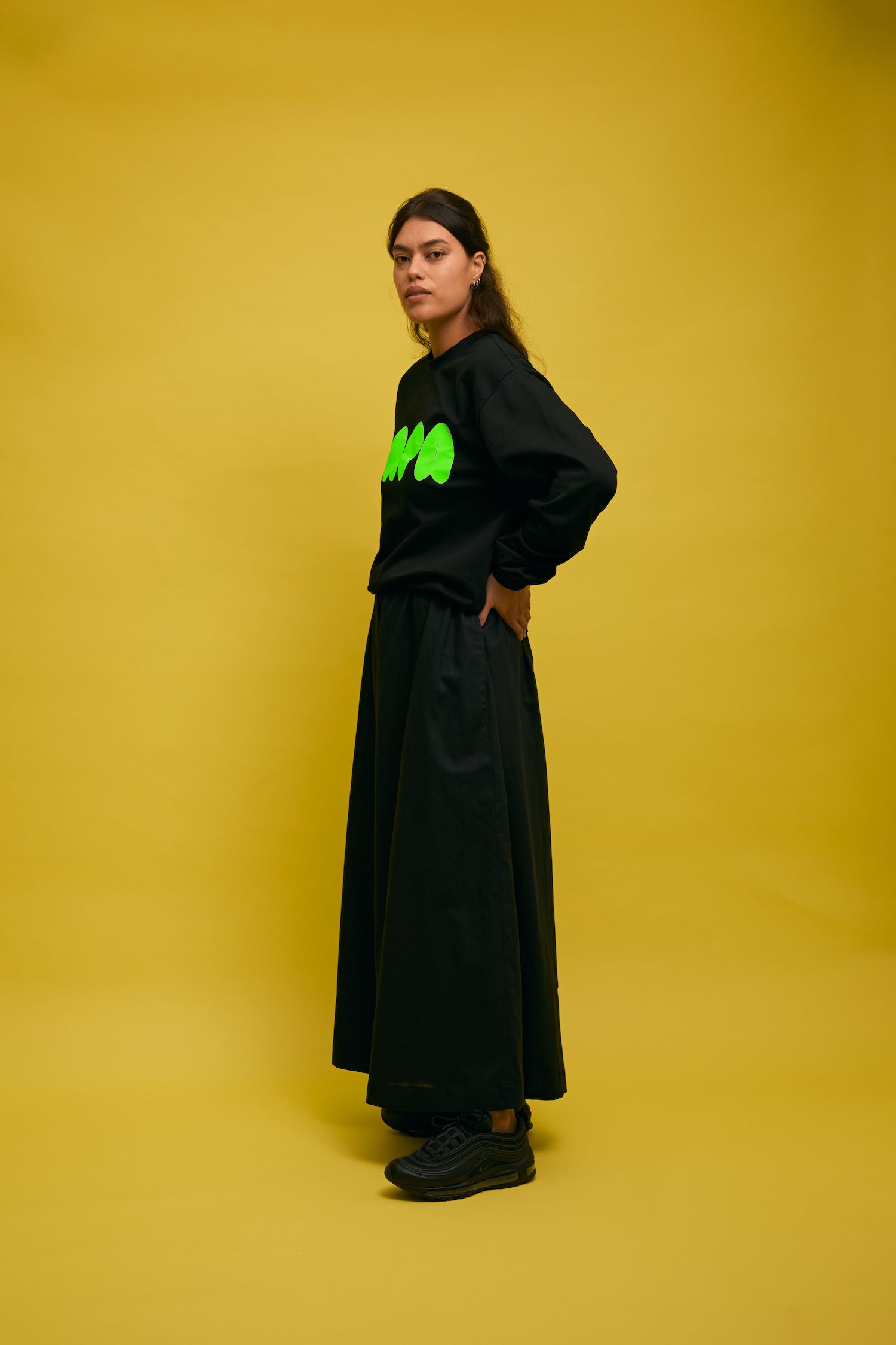 Karen Valerie standing in front of yellow wall wearing black AS colour longsleeve tee with green PAPA text and green hibiscus print