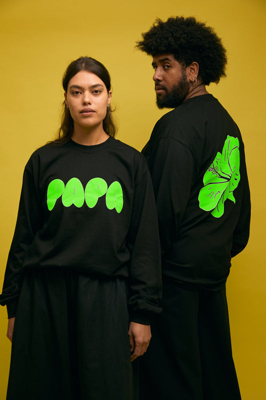 Karen Valerie and Peter Wing standing in front of yellow wall wearing black AS colour longsleeve tee with green PAPA text and green hibiscus print