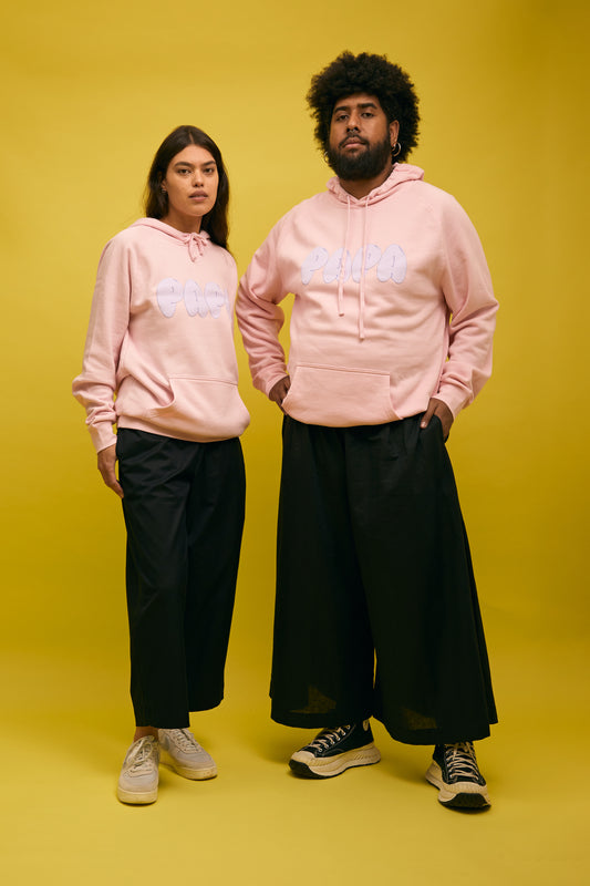 Karen Valerie and Peter Wing standing in front of yellow wall wearing pink AS colour hoodie with Papa text on front and Hibiscus on the back