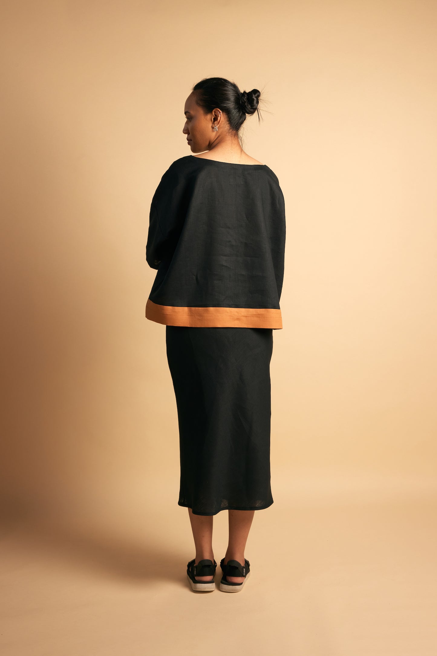 Tongan woman, girl standing in front of beige wall wearing black linen top with orange hem and matching black linen bias cut skirt